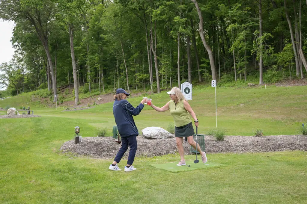 Two women smiling and relaxing after a round of park golf at 1Club in Vernon, NJ, with golf clubs and scenic greenery in the background.