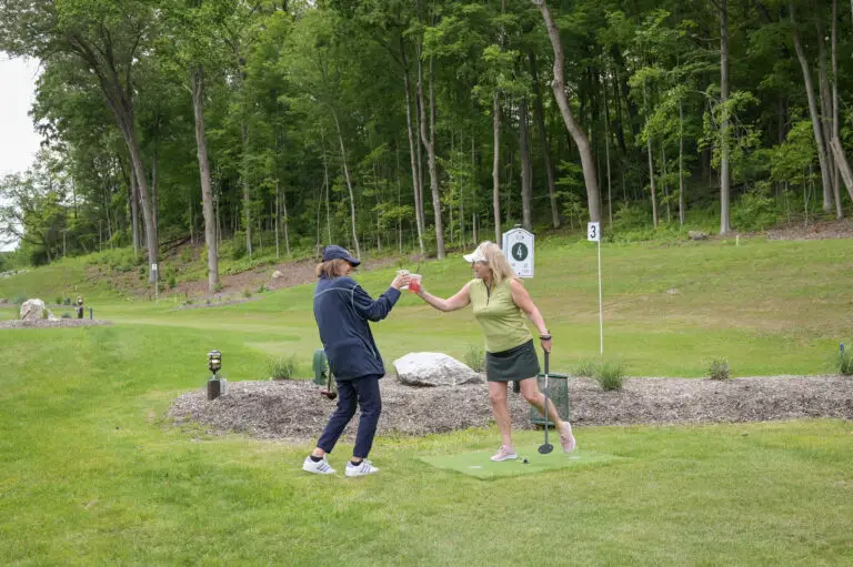 Two women smiling and relaxing after a round of park golf at 1Club in Vernon, NJ, with golf clubs and scenic greenery in the background.