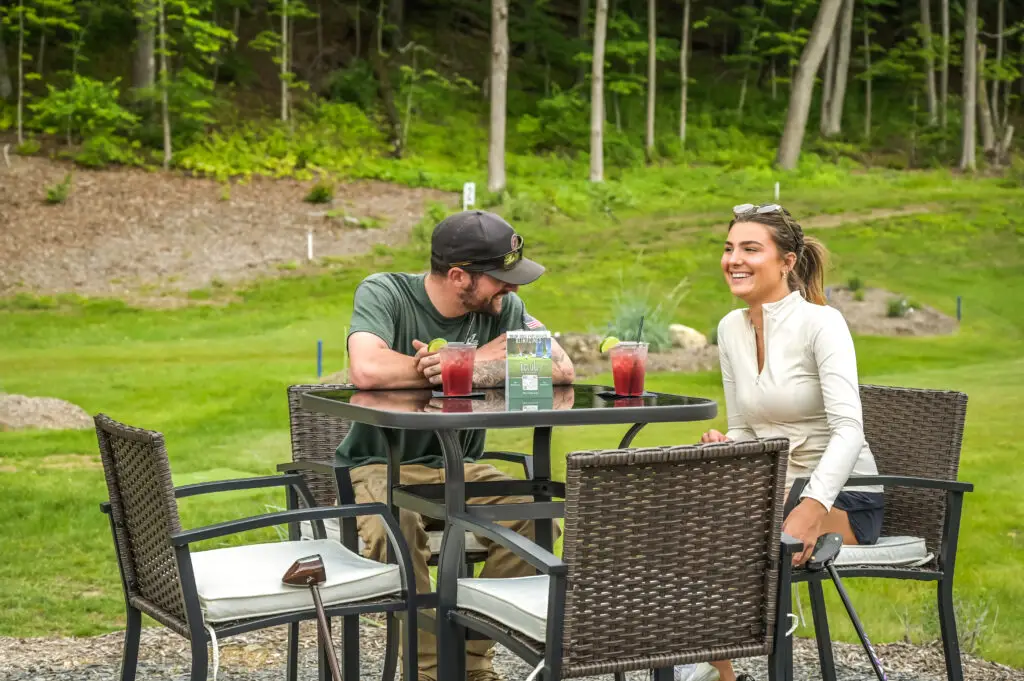 Couple sitting outdoors at 1Club, drinking and smiling with the park golf course visible in the background.