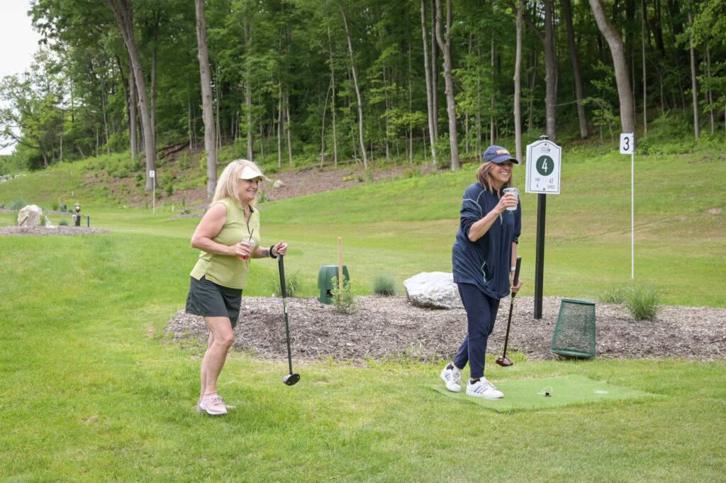 Two women having fun while playing park golf at 1Club in Vernon, NJ, laughing and enjoying the game on the scenic course surrounded by lush greenery.